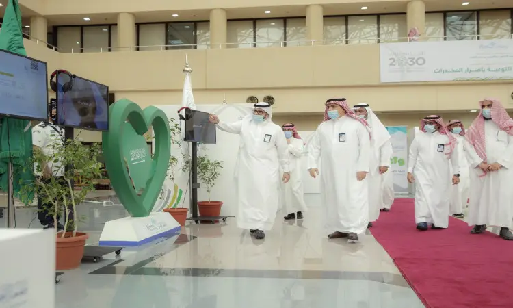 مجموعة من الرجال الثياب البيضاء، ويضعون الغترة أو الشماغ السعودي مع العقال، وبعضهم يرتدون البشت السعودي، يسيرون في الداخل بالقرب من زينة خضراء على شكل قلوب ونباتات مزروعة في أصص وشاشة عرض كبيرة، وذلك ضمن فعاليات معرض "واعي" للتوعية بأضرار المخدرات في جامعة القصيم. يُلاحظ أن بعض الحضور للكمامات، كما تلاحظون وسجادة ملونة ذات عنابي في الخلفية.