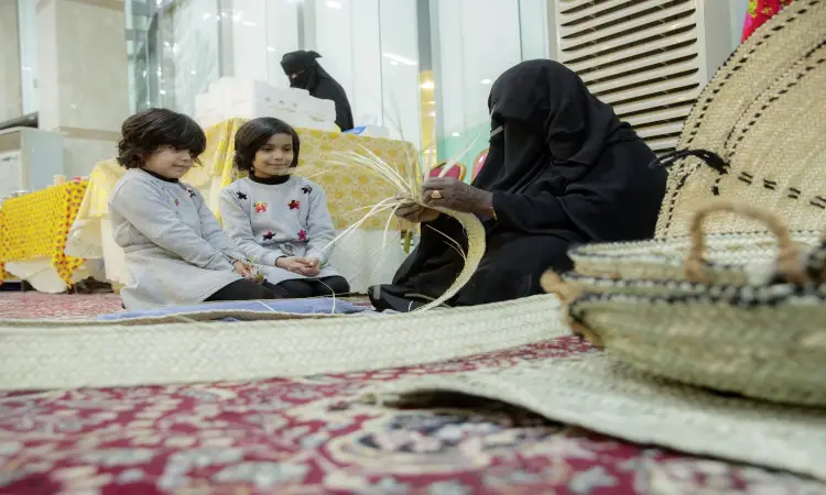 امرأة مسنة ترتدي ملابس بيضاء وبشتاً سعودياً، تجلس على سجادة مزخرفة وهي تنهمك في خياطة حصير من السعف. بجانبها، ملابس فلوريدا فتاتانتان الصغيرة بهن التقليدية وهن تتابعن باهتمام وإعجاب الكبارين خطواتها المتقنة، فيما ترتدي إحداهن الشماغ السعودي وتضع الأخرى الغترة السعودية مع عقال فوق الرسائلهن. تثتر حول هن سلال من السعف، في مشهد يعكس الاعتزاز بالهوية الوطنية ويجسد قيم النبيلة التي تؤكد عليها حملة سمو أمير منطقة القصيم.