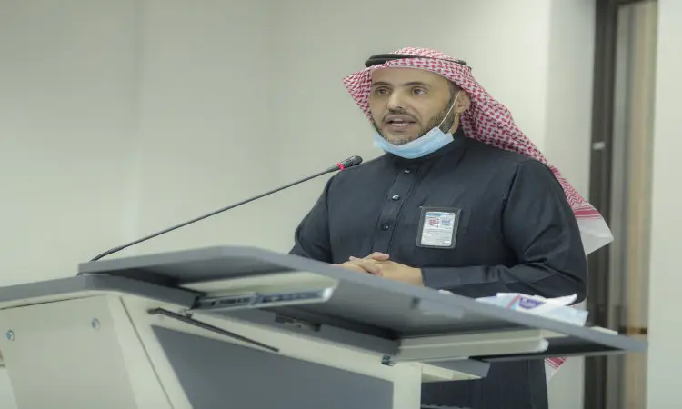 رجل مرتدياً مشهوراً وبشتاً سعودياً، مع شماغ سعودي وعازف ميكروفون، محترفاً من على منصة ب خلال ورشة عمل في كلية الاقتصاد والإدارة في القصيم. تظهر على بطاقته التعريفية، بينما يرتدي كمامة أسفل الصدر للرجال. واخيرا ذلك في اعتبارات الاعتبارات الداخلية الأخلاقية للفعالية الأكاديمية.