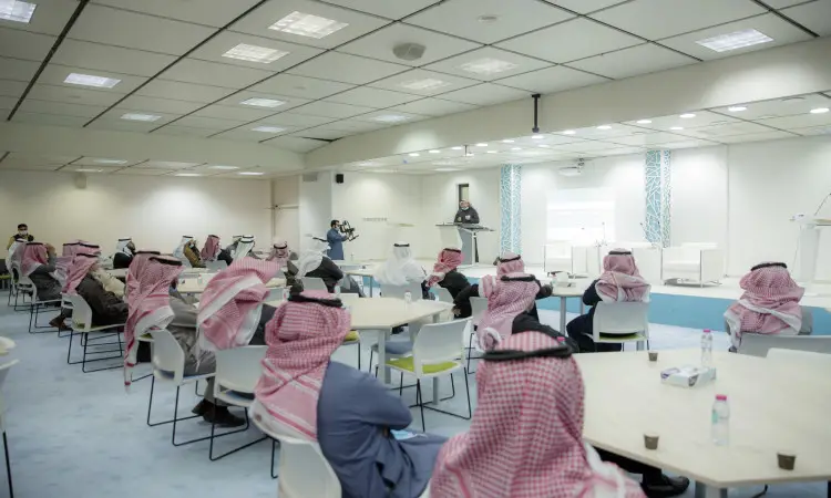 يجتمع مجموعة من الرجال مرتدين الغترة أو الشماغ والعقال، وقد أكملت إطلالته بالبشت السعودي، حول طاولات متجمعة في قاعة مؤتمرات واسعة حديثة بكلية الاقتصاد والإدارة في جامعة القصيم، حيث يشاركون في ورشة عمل متخصصة واختراع ويستمعون باهتمام لمتحدث يلقي كلمته على المنصة.
