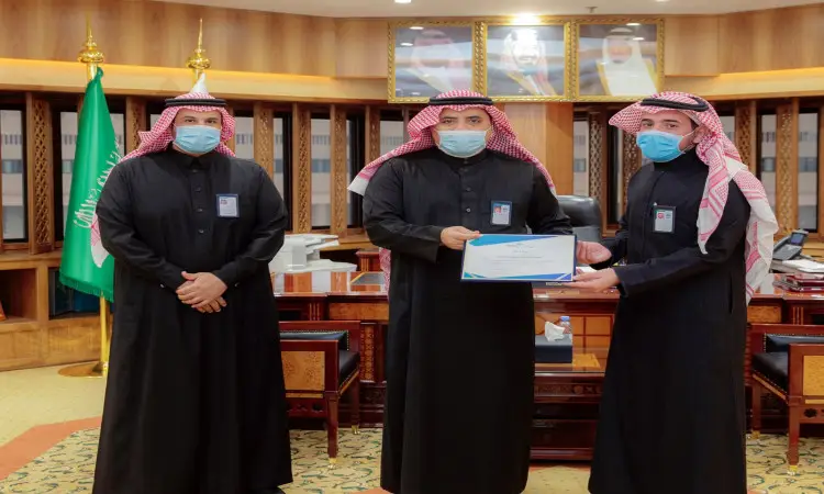 التزام ثلاثة رجال بالغتة والشماع والعقال والبشت السعودي في أحد مكاتب جامعة القصيم، ملتزمين بارتداء الكمامات. هو أحد بتسليم حصل على تقدير احتفاء بإنجاز فريق بحثي للحصول على الميدالية الذهبية في مؤتمر ايتكس2021. للأخذ في الاعتبار لوحة تحمل العلم السعودي والأقل من مساهمة الأسرة بإطار رسمي.