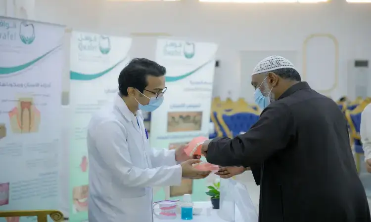 بدلة طبيب يرتدي المعطف الأبيض الناصح السليمة بالأسنان باستخدام نموذج يي أمام رجل يرتدي الشماغ السعودي مع العقال والبشت، وذلك في جناح تعريفي ينظمه قوافل طب الأسنان في القصيم، حيث يحفظ في الخلفية ملصقات توعوية داخل مركز قبة.