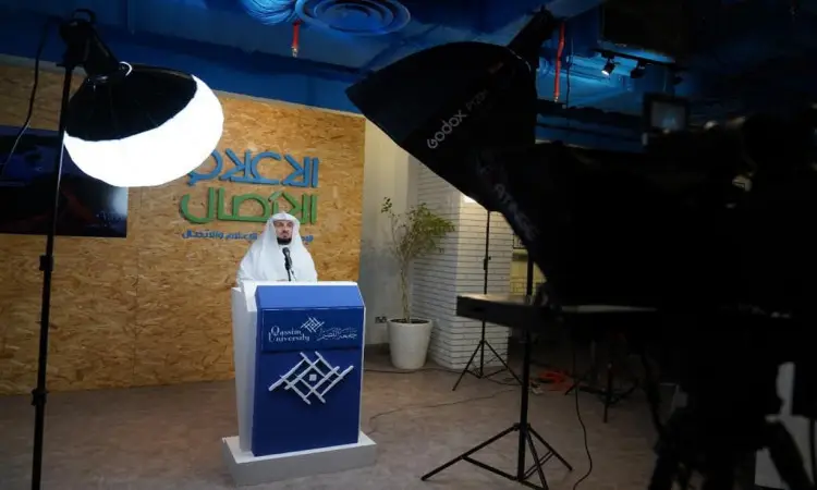 رجل الاتصال مرتدياً غترة أو شماغاً سعودياً وقالاً وبشتاً سعودياً أمام منصة الإسبانية داخل الاستوديو مُجهّز بأضواء رأسية وكاميرات تصوير، مشاركًا في أحد برامج جامعة القصيم. احفظ خلفه شاشة تحمل كتبًا باللغة العربية وشعار الجامعة، بالإضافة إلى إشارات ذات صلة ببقية الدوائر الحكومية.