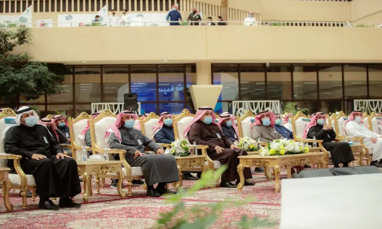 جلسة عدد من الرجال مرتدين الغترة أو الشماغ والعقال والبشتاء السعودي، مع وضع الكمامات الطبية، على مقاعد فخمة داخل قاعة مزينة بالأزهار والسجاد خلال فعالية يوم الطالب في القصيم.