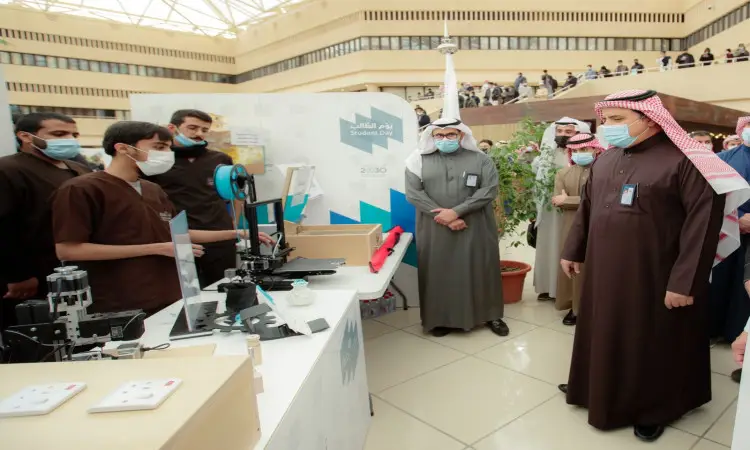 عدد من الرجال، من بينهم رؤساء الجامعة وأعضاء الأندية الطلابية، وظهور الشماغ السعودي أو الغترة والعقال والبشت السعودي، ويضع التعاون مع الكمامات الطبية، يتابعون باهتمام شابًا يقوم بإظهار عملي لطابعة ثلاثية الأبعاد وذلك ضمن معرض داخلي يضم لوحات تعريفية ونباتات زينة.