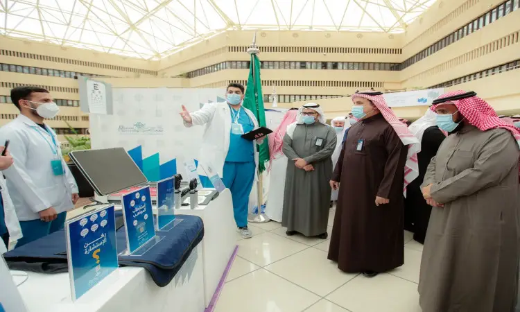 ألت مجموعة من الرجال مرتدين الغترة أو الشماغ والعقال، إضافة إلى البشت السعودي، حول جناح معرض طبي ضمن فعاليات يوم الطالب في جامعة القصيم. ويستمع الحضور باهتمام إلى شرحه أحد المختصين الصحيين حول الأجهزة الطبية، وذلك داخل القاعة تغطيها سقوف مضيئة.