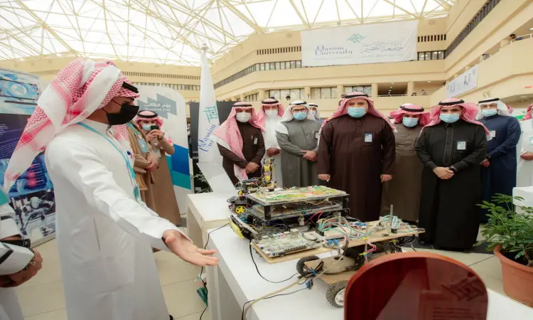 تويوتا مجموعة من الرجال تولى الغترة أو الشماغ والعقال مع البشت السعودي، وعليهم متعة رئيس الجامعة، أحد العارضين وهو يشرح مشروعًا إلكترونيًا، وذلك ضمن معرض داخلي للأندية الطلابية في يوم الطالب في القصيم، حيث تُزين اللافتات أروقة المعرض وتعتمد تدابير الاحترازية بارتداء الكمامات.