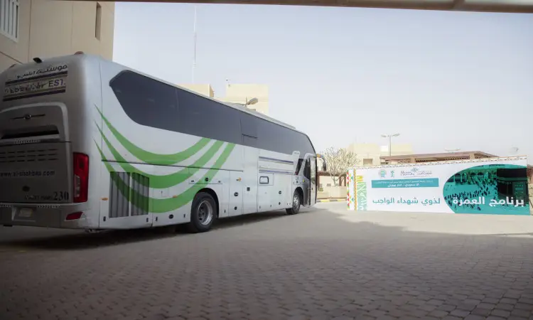 A silver and green color near the prominent Arabic-language Nomek program, possibly related to Qassim or the Umrah program, has stopped playing. Review in a series of a number of people wearing the Saudi Ghutra or Shemagh with Eqal, as well as the Saudi Bisht, giving the venue a feel appropriate for a university event. An area paved with geometric tiles and surrounded by modern buildings in the background under a clear sky.