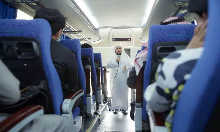 A man dressed in traditional Saudi attire, represented by a Saudi Ghutrah or Shemagh with a Saudi headband and besht, takes the microphone in a well-lit track to provide directions and guidance to participants in the Umrah program from Qassim University. Those sitting on the blue explanations listen attentively, and among them are the families of the martyrs.