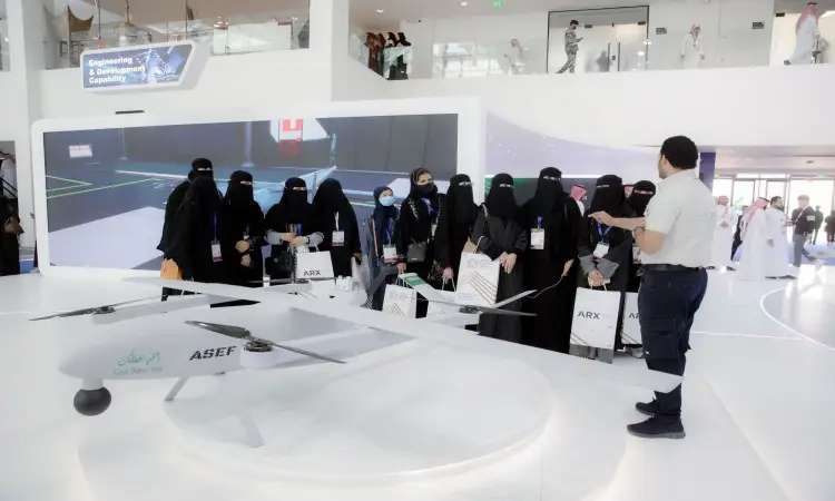 A Florida man leads a group of female students, dressed in black abayas and niqabs, at the International Defense Exhibition in Riyadh, wearing the Saudi Ghutrah, Eqal and white Saudi bisht. They listen intently as he shows them a model drone and a screen in one of the state-of-the-art drone halls for display.