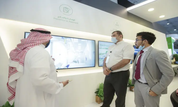 At the "Prevention and Treatment" pavilion during the Middle East Poultry Exhibition in Riyadh, three men exchange pleasantries; two of them are dressed in long-sleeved clothing, while a third is wearing a Saudi tunic and men's shirt with an official university certificate including a national ID. Two of the attendees are wearing masks while parked in front of a large projection screen at the booth.