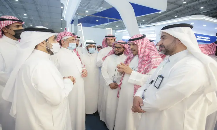 A group of men dressed in traditional Saudi attire, including the thobe, shemagh or Saudi ghutra with an aqal, as well as the Saudi besht, gather and chat at the Middle East Poultry Exhibition in Riyadh. They are wearing masks and ID cards, while the exhibition pavilions are visible in the background.