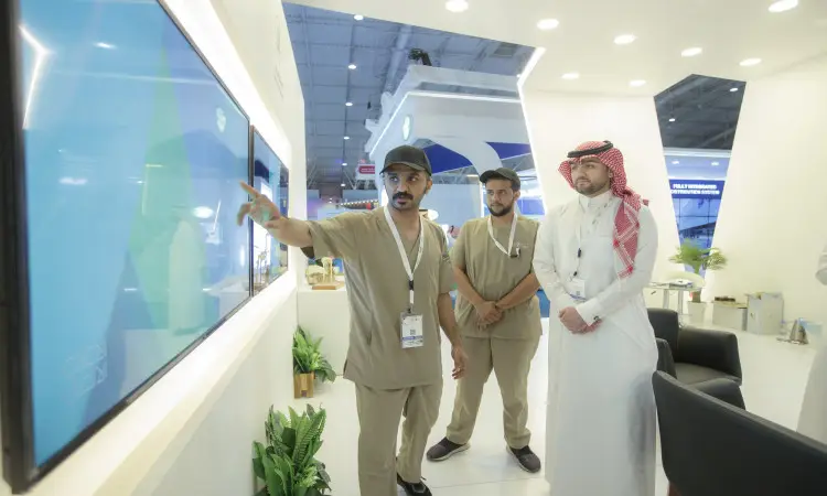 Three men gather in front of a booth at the Middle East Poultry Exhibition in Riyadh, one wearing a Saudi bisht with a white thobe, Saudi hijab and aqal, while the other two are dressed in beige-green uniforms. One of them points to a large interactive screen while the others watch intently as part of Qassim University's participation in the prestigious event.