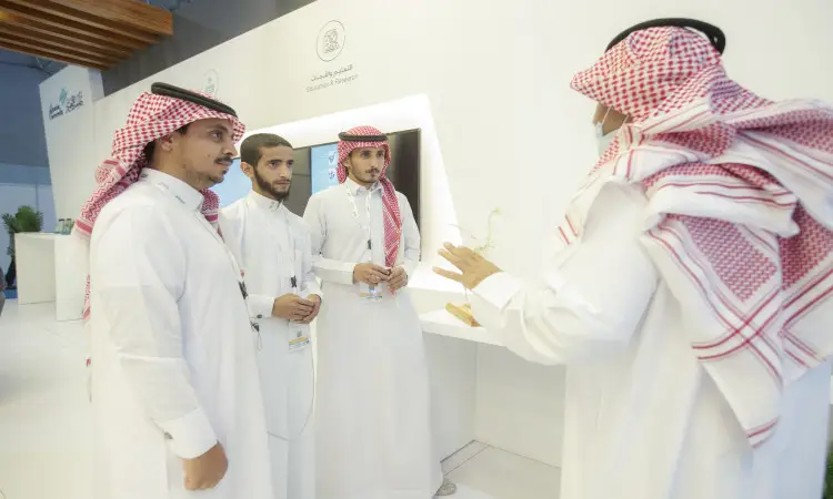 Four men in traditional Saudi dress, including a ghutra or shemagh and a headband, chatting inside a modern auditorium in Riyadh. Three of them pay attention to the fourth, who often animates the gestures while explaining. She has successfully whitewashed items from the Middle East Poultry Exhibition.