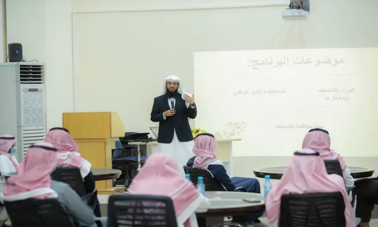 A man dressed in the official Saudi uniform, consisting of a Ghutra or Shemagh and Eqal in addition to a Saudi Basht, gives a presentation on the topic of intellectual fortification to a group of men wearing Ghutra or Shemagh and Eqal sitting around round tables inside the auditorium. The lecturer controls the screen with a text in Arabic to address the topic of "diagnosis."