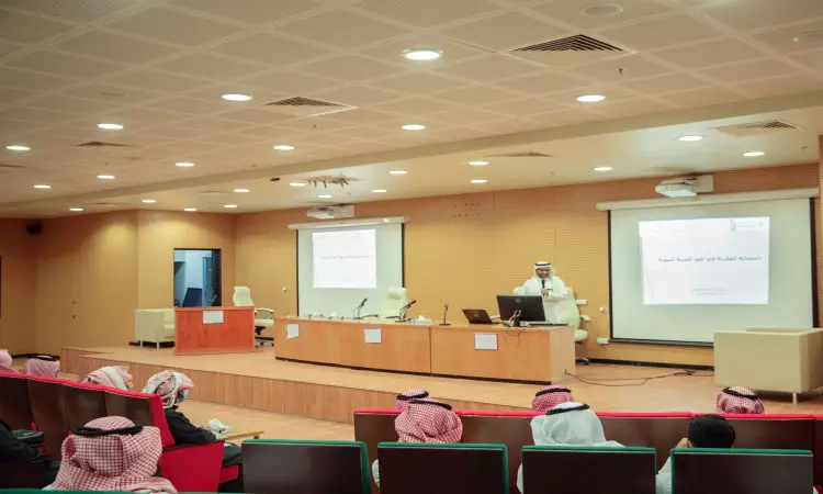 A man behind the podium in a conference hall in Qassim, giving a presentation on the Intellectual Fortification Program, while two screens are filled with Arabic texts that adhere to this program. A number of attendees dressed in Saudi shemagh, or Saudi ghutra, aqal and bisht, sit listening intently to a presentation on any occasion of the university's prestige.