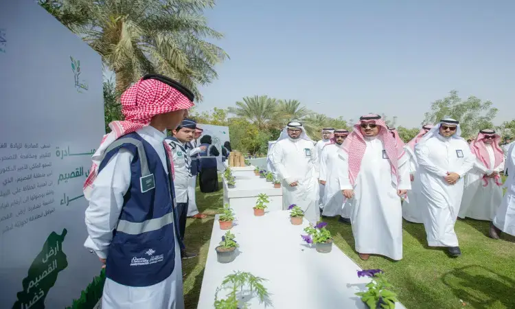 A number of men wearing the official Saudi uniform, including the Ghutra or Shemagh with a square of the Saudi Aqal and Bisht, as well as one participant in his official jacket, gathered outside around tables with small seedlings on them. This was part of the tree planting efforts that took place under the umbrella of Al Nakheel Company and next to a sign bearing the words "Qassim Green Land" in Arabic, under the auspices of Qassim University's economic sustainability program.