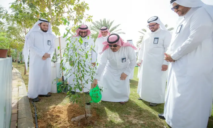 A group of men wearing the Saudi Arabian ghutra or shemagh, headband and bisht gather outdoors as one of them waters a sapling using a green irrigation breaker, in support of the greening of Qassim, on a sunny day as part of the Qassim University events.