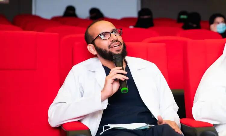 يرتدي الرجل سترة بيضاء ونظارة طبية، ويضع على شماغاً سعودياً وقالاً، بالإضافة إلى بشت سعودي، يُمسك اً أثناء الجلوس على الكرسي الأحمر في القاعة الكبرى بكلية طب الأسنان في جامعة القصيم، ويرتفع بنظره للأعلى. يؤخذ في الاعتبار عدد من الحضور حيث يحضرون كميات أو أعضاء.