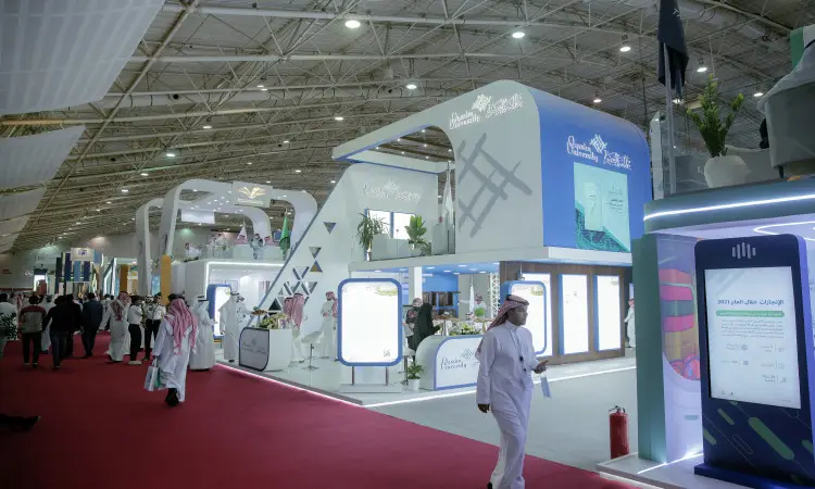 يتجول الزوار في قاعة العرض الحديثة ضمن فعاليات المعرض الدولي للتدريب في الرياض، حيث تمثل الأجنحة البيضاء والشاشات الرقمية ويغطي سجادة الطاولة باللون الأحمر، وتبرز اللافتات العربية بشكل واضح. ويظهر بعض الحضور وهم بحضور الشماغ السعودي أو الغترة السعودية مع العقال والبشت السعودي التقليدي، مما يعكس الهوية الوطنية. وقد أخذت القاعة بإضاءة مشرقة ونباتات خضراء في أرجائها لتضفي بشكل مريح وراقية تليق بالتعاون مع جامعة القصيم ومكانتها الشهيرة.