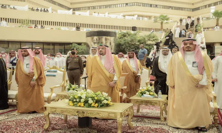 A group of men dressed in a ghutra or shemagh and aqqal, wearing the official Saudi bisht, called a luxurious hall with wooden chairs, flower arrangements and a red carpet, for Qassim University's "Innovator Showcase", where creative students and event guests, including a number of dignitaries in their uniforms, gather in a capital of trust and prestige.