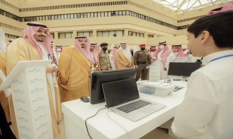 A group of men wearing the Saudi Ghutra or Shemagh, Egal and Bisht, accompanied by security members, gather around a touch platform with computers, interacting with a young man during the activities of the Innovator 2022 exhibition organized by Qassim University in one of the indoor halls.