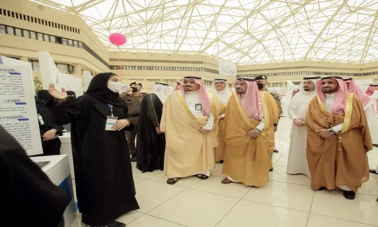 A number of men wearing a ghutra, shemagh, aqal, and Saudi bisht gather to listen to a woman in white giving a presentation as part of a glass-engineered indoor exhibit during the Innovator 2022 event at Qassim University.