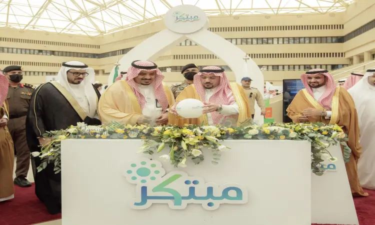 A number of men dressed in a Saudi Arabian ghutra or shemagh with an aqal, in addition to the traditional Saudi bisht, gathered around a display stand with bouquets of flowers and the "Innovator" logo as part of the Innovator 2022 exhibition at Qassim University. A coordinated presence under the high glass ceiling illuminated the background, including the official and distinctive signature of the show.
