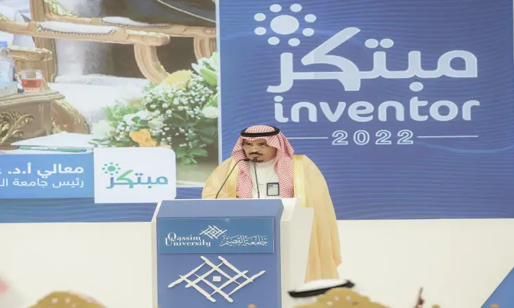 A man dressed in a Saudi Arabian ghutra, headband and bisht gives a speech from the stage during the Innovator 2022 exhibition at Qassim University. In the background, a banner in Arabic and English celebrates innovative students.
