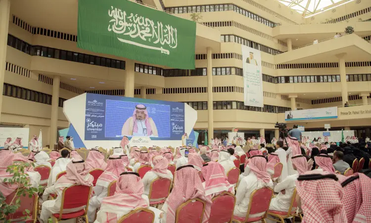 A large gathering of men wearing the Saudi Ghutra or Shemagh, along with the Saudi Aqal and Bisht, are participating in an official conference being held inside Qassim University. Finally on stage, a large man, the Emir of Qassim, delivers his speech in the presence of the Saudi flag and a large screen behind him whose image is projected as part of this official ceremony.