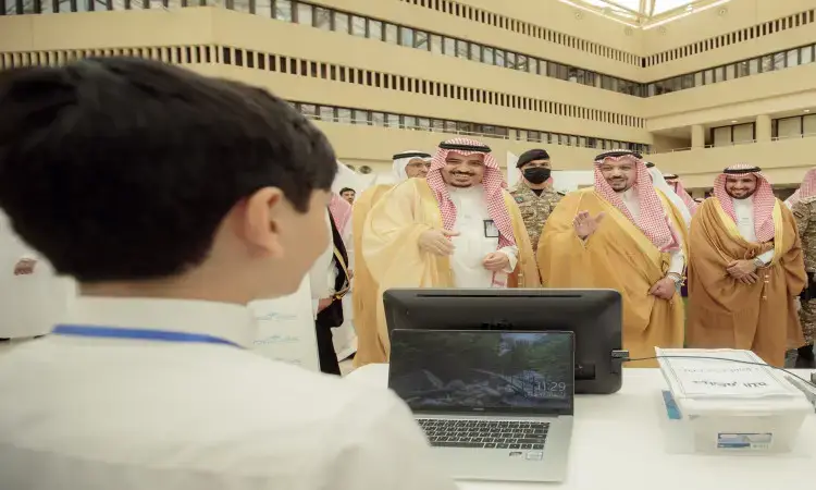 A group of Saudi men, dressed in Saudi Arabian ghutra, shemagh, aqal and bisht, smile and interact with a child in front of a computer screen and paperwork inside a modern building, during the Creators' Fair at Qassim University.