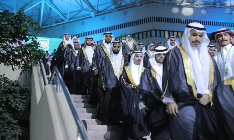 مجموعة من الرجال تولي الزي السعودي التقليدي، المكوّن من الغترة أو الشماغ، والعقال، والبشتاء السعودي، يتقدمهم صاحب السمو أمير منطقة القصيم، ينزلون السُلَّم في جامعة القصيم خلال تخريج الطلاب، وذلك تحت إضاءة مشرق وسقف هندسي.
