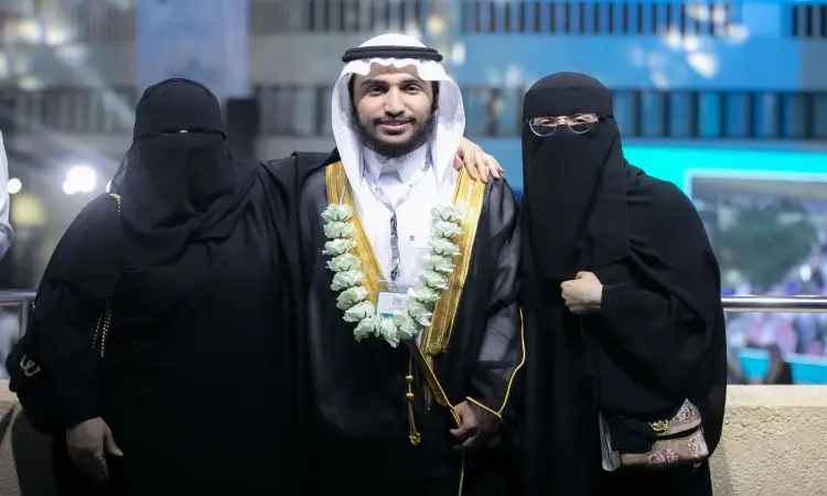 يلتقي ثلاثة بما في الصورة خلال تخريج الدفعة التاسعة عشرة في القصيم: يظهر رجل يرتدي غترة أو شماغًا سعوديًا مع عقال وبشت سعودي، وقد زُين بعقدة سوداء من الورود، يتوسط سيدتين يُحتمل أن تكون وتدتيه، ترتديان أت تحمل ونقابًا. التُقطت الصورة في الهواء الطلق للإضاءة ومبنى الجامعة.