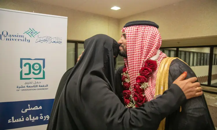 يرتدي رجل سعودي اللباس الأصلي، يشمل الغترة أو الشماغ والعقال والبشت السعودي، ويرتدي طوقاً أسود من الزهرة الحمراء حول عنقه ويحتضن والدته التي ترتدي العباءة بجانب لوحة تحمل شعار جامعة القصيم، وذلك من خلال تحرير للدفعة التاسعة عشرة.