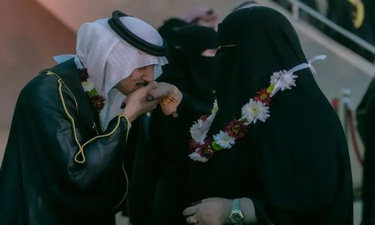 خلال حفل تخرج الفوج التاسع عشر بجامعة القصيم، شوهد رجل يرتدي البشت السعودي التقليدي والغترة والعقال وهو يُقبّل يد امرأة ترتدي عباءة سوداء ونقابًا، وكلاهما مُزيّن بأكاليل الزهور. تظهر شخصيات مُشوّشة في الخلفية، مُسلّطةً الضوء على هذه اللحظة المؤثرة التي تشاركتها الأمهات في هذه المناسبة المُميّزة.