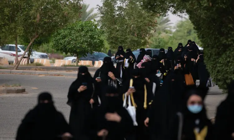 مجموعة من طالبات الدفعة التاسعة عشرة وهن يرتدين العباءة السوداء والبشت السعودي، ويسدلن الغترة السعودية فوق رؤوسهن، يتقدمن على ممر تصطف أمامه على جانبيه داخل حرم جامعة القصيم، بينما يستخدم بعضهنهنهن. لأخذ في الاعتبار سيارات متوقفة.