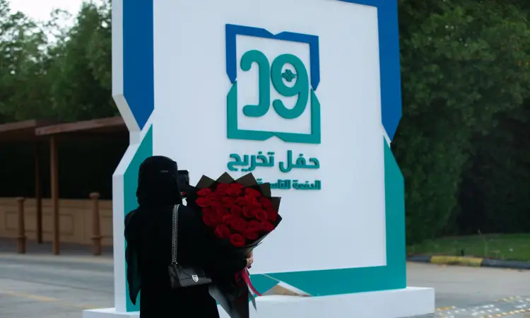 امرأة ترتدي سترة وبشتاً سعودياً، وتضع غترة وعقالاً، تحمل باقة كبيرة من الورود الحمراء وتقف بجوار لوحة تحمل الرقم 29 ونصاً عربياً يحتفي بحفلات تحرر لطالبات الدفعة ١٩ في جامعة القصيم. طبخ قطع والمرات في الخلفية.