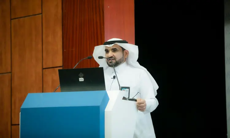 يرتدي رجل غترة سعودية وقالاً وبشتاً تقليدياً، غالباً من على منصة العرض خلال ورشة عمل للتغيير إلى الفصول الثلاثة في جامعة القصيم؛ ممسكًا بجهاز تحكم عن بعد، وأمامه جهاز كمبيوتر محمول وخلفيه جدار بانِل خشبي.