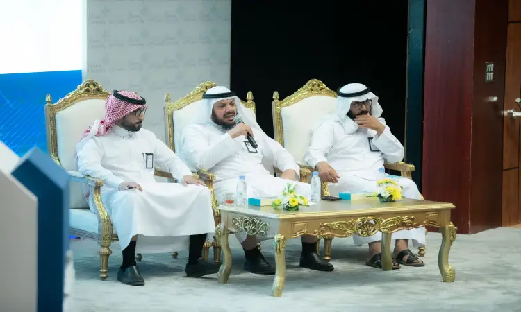 يجلس ثلاثة رجال مرتدين الغترة أو الشماغ والعقال مع البشت السعودي على كراسٍ فخمة خلف طاولة مزينة بالزهور وقوارير المياه، مشاركين في حلقة نقاشية حول تأسيس التأسيس ضمن ورشة عمل أو مؤتمر شعبي عُقد في جامعة القصيم.