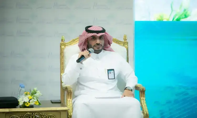 رجل جلسة مرتدياً الغترة السعودية والعقال والبشت السعودي كرسي مزخرف بإطار ذهبي في جامعة القصيم، ممسكًا بميكروفون أثناء المشاركة في ورشة عمل تناقش نظام الفصول الثلاثة. وقد وضعت وثائق عليه، وتوجد إلى جواره زجاجة بعض الماء، في حين تظهر خلفه شاشة زرقاء.
