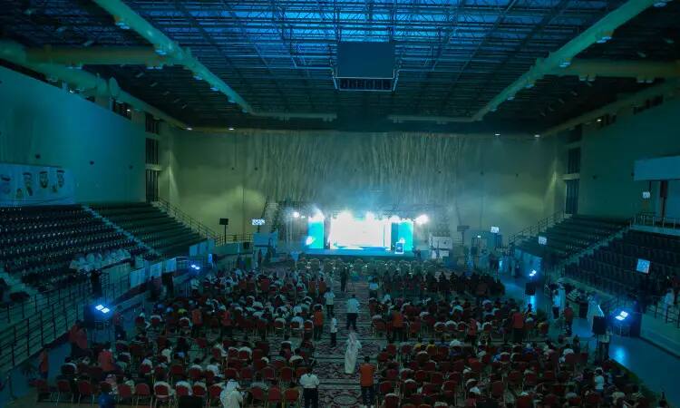 A large auditorium at Qassim University is lined with rows of red chairs, filled with attendees facing a brightly lit stage. Seated at the front are the distinguished students, dressed in the official Saudi uniform of shemagh, akal and bisht, while the blue light creates a majestic atmosphere under high ceilings and empty side bleachers.