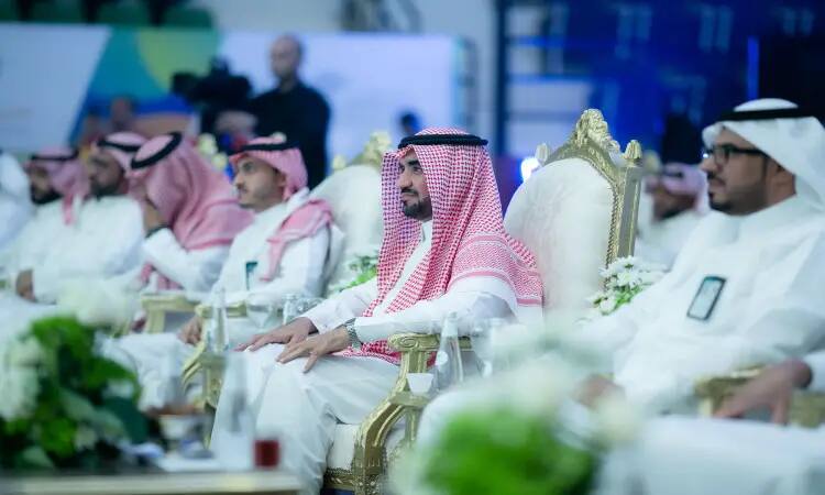 A group of men dressed in Saudi shemagh, aqal and besht sit on plush chairs during a meeting of events organized by Qassim University or its summer club. The tables in front of them are adorned with formations and vases, vaguely suggesting a formal atmosphere in which outstanding students are being honored.