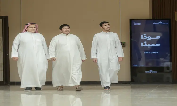 Three students, dressed in a fourth outfit with a Saudi Arabian ghutra and a qalal, walk into a modern building with a shiny floor. Behind them, a digital screen displays a text in Arabic, depicting a sad scene of children at Qassim University at the start of the university. It looks like one of the students might spruce up his formal look by wearing a Saudi besht.