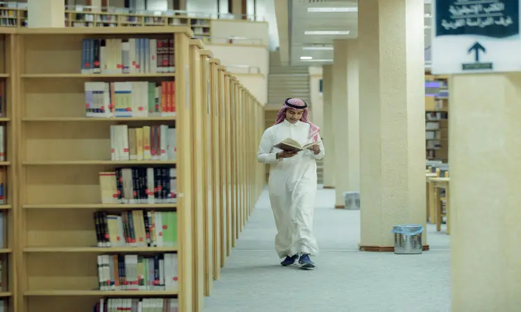 A man wearing a Saudi Arabian ghutra and traditional bisht reads a book as he passes between bookshelves on a sidewalk in the library at Qassim University. To make a decision in an internal staircase gives the surprise of an academic official.