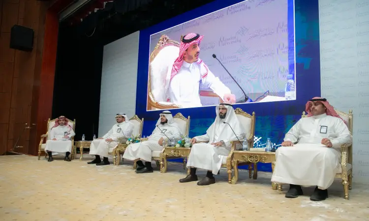 مجموعة خمسة رجال يرتدون الغترة أو الشماغ والعقال والبشتاء السعودي على كراسٍ مزخرفة فوق المنصة، بينما الشتاء أحد في الميكروفون خلال فعاليات برنامج طموح. وظهرت الشاشة الكبيرة صورة رئيس الجامعة، مما يضفي على هذا الحدث الرسمي وترحيباً رسمياً للطلاب في جامعة القصيم.