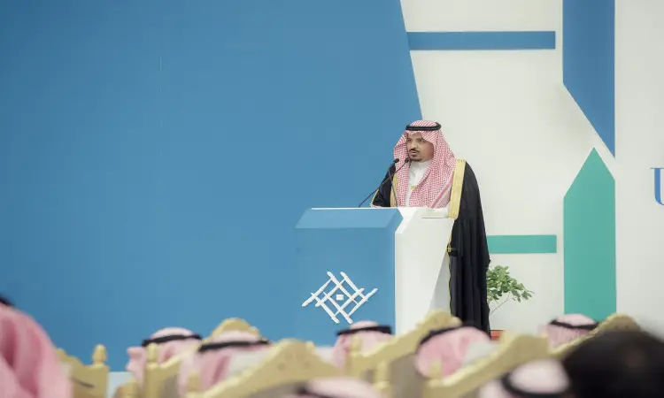 رجل القط مرتدياً البشت السعودي والغترة والعقال على منصة الخطابة عبر جامعة القصيم بمناسبة اليوم الوطني الثاني والتسعين. ويستمتع الحضور، الذين تأثروا بالشماغ والعقال والأزياء السعودية التقليدية، باهتمام لكلمته. فكر في تصميمات هندسية باللون الأزرق والأبيض تضفي طابعًا رسميًا ومميزًا على الفعالية.