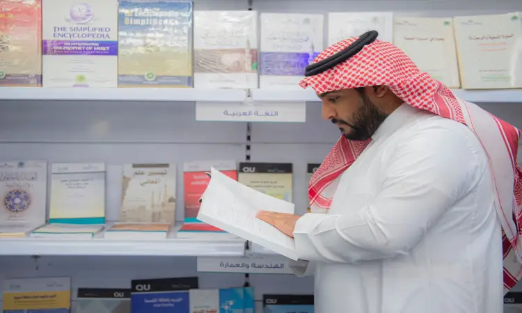 رجل يرتدي الزي السعودي الرسمي، بما في ذلك الشماغ السعودي والعقال والبشت، نسخة كتابية خلال معرض الرياض الدولي للكتاب. يؤمن خلفه رفوف تحمل كتبا متنوعة باللغتين العربية والإنجليزية في مجالات متعددة.