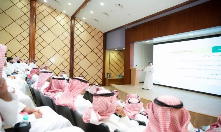 Dressed in traditional Saudi attire, consisting of a ghutra or shemagh, aqal, and Saudi besht, a workshop on energy construction is presented to a number of government-represented attendees. The meeting is held in a lecture hall in Qassim, where attendees sit on the ecosystem while Arabic-language texts related to the workshop's themes are projected on a large screen.