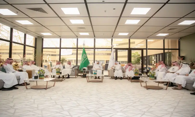 A number of men dressed in the Ghutra or Shemagh and Eqbal, some in the official Saudi winter dress, are gathered in a large modern hall with large windows. Saudi Arabia's flags adorn the hall and flowers are placed on the tables as they hold key discussions as part of the Qassim government workshop on energy conservation.
