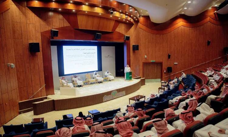 في قاعة مؤتمرات واسعة ذات جدران خشبية، حضر جمع من الرجال مرتدين التحول الغترة السعودية أو الشماغ والعقال، بالإضافة إلى البشت السعودي، فعالية جامعية نظمَتها جامعة القصيم حول موضوع في التعليم. وقف العرض على الشاشة الرئيسية مخاطبًا الحضور الرسمي، بينما عُرضت على الشاشة الرئيسية تقديم خاصة بمناسبة اليوم العالمي للمعلم.